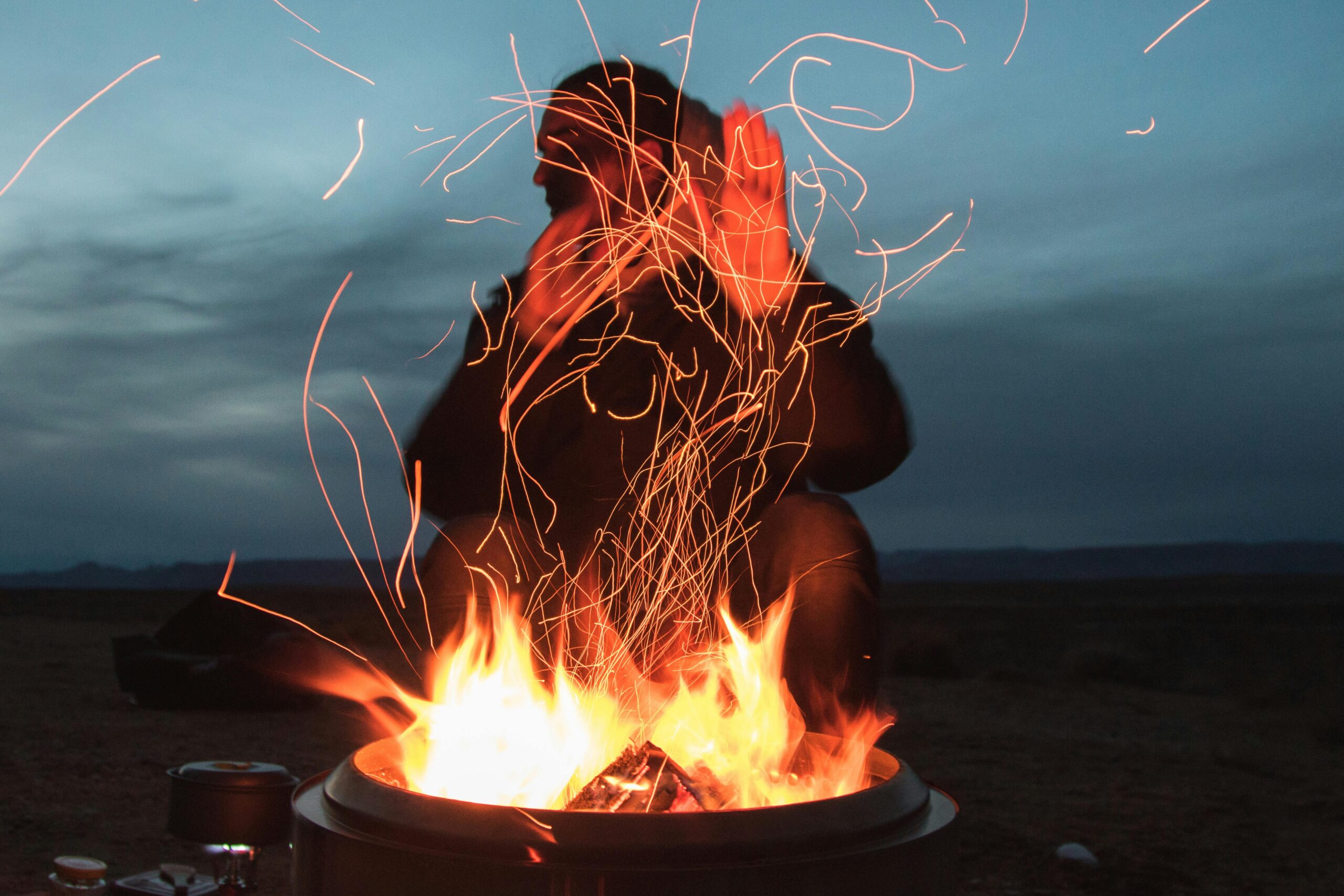 A person enjoying warmth by a vibrant campfire at twilight, sparks flying into the night sky.